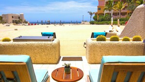 A beachside seating area with a view of the ocean, a palm tree, and a building in the background. - Terrasol Oceanview Condo with Patio (Cabo San Lucas)