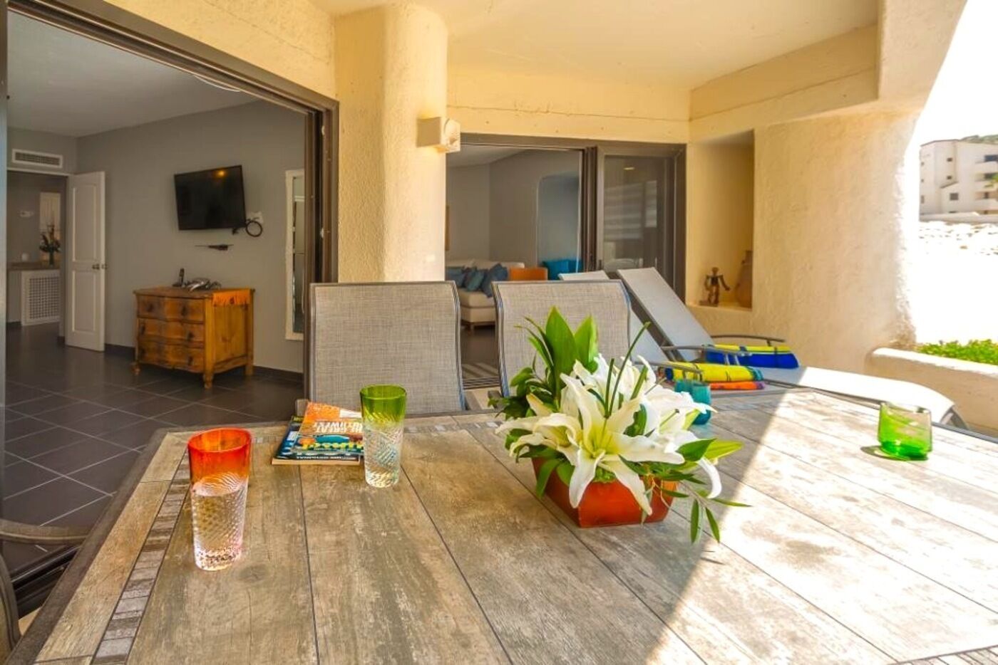 A sunlit patio with a wooden table, a potted plant, and a television in the background.