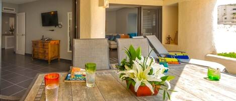 A sunlit patio with a wooden table, a potted plant, and a television in the background.