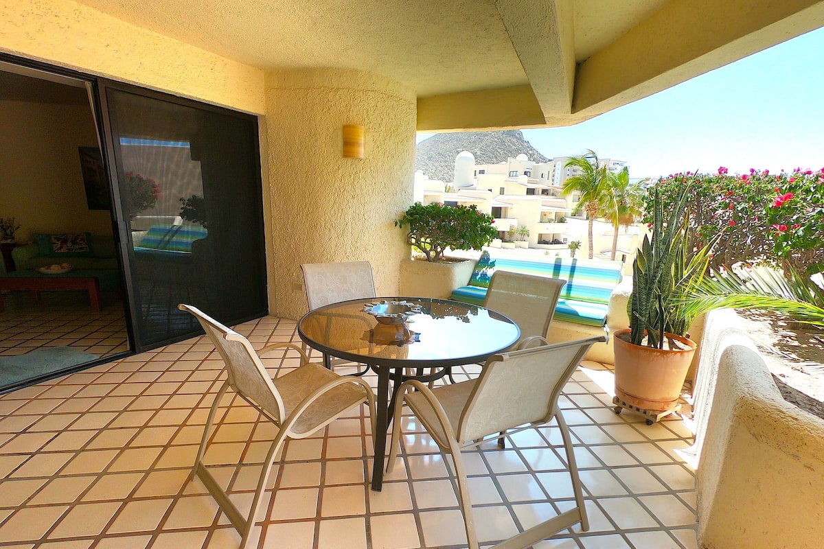 Eine Terrasse mit rundem Tisch, vier Stühlen, einem Blumentopf und Blick auf ein Gebäude sowie einen Pool im Hintergrund.