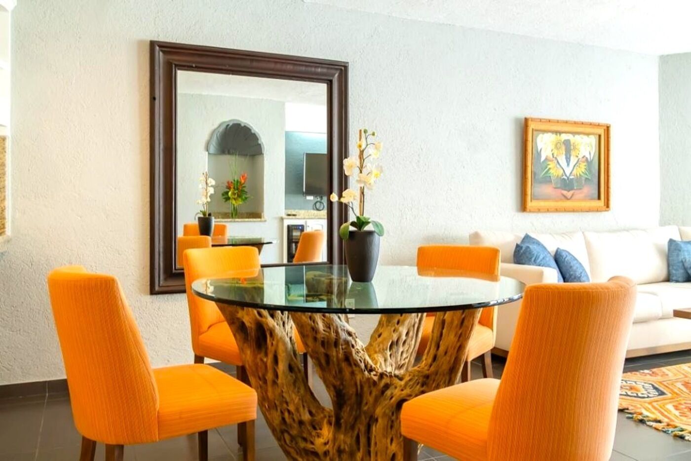 Un comedor con una mesa de vidrio y sillas naranjas, un espejo grande y un cuadro en la pared.