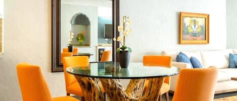 Un comedor con una mesa de vidrio y sillas naranjas, un espejo grande y un cuadro en la pared.