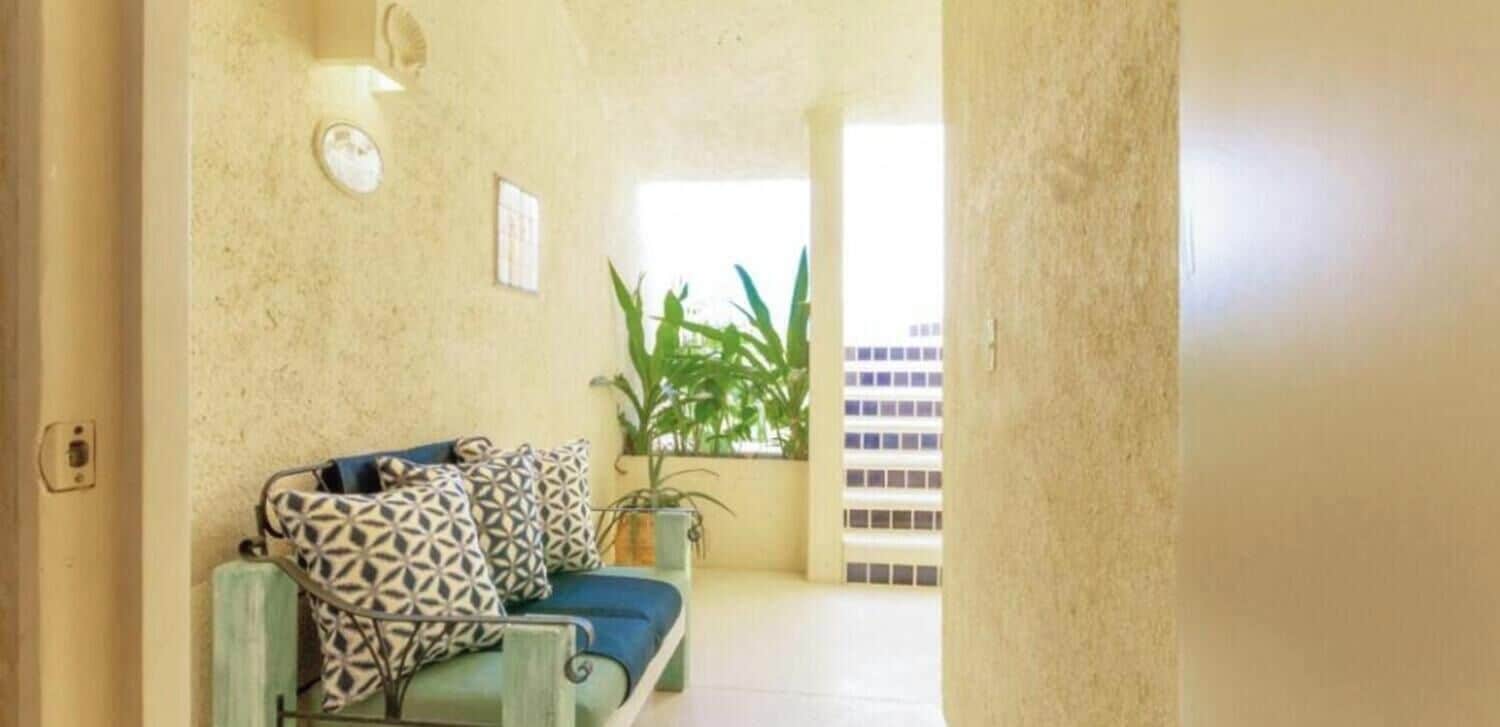 Uma sala de estar acolhedora com um sofá e almofadas estampadas, uma planta em vaso e vista para um edifício através de uma janela.