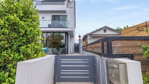 A modern two-story building with a gated entrance, a balcony, and a well-maintained hedge. - @ Marbella Lane - Luxe 4BR Haven w/ Skyscape Views (Huntington Beach)