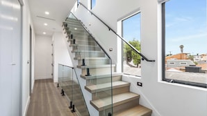 A modern staircase with glass railings and wooden steps, leading to a balcony with a view of the city. - @ Marbella Lane - Luxe 4BR Haven w/ Skyscape Views (Huntington Beach)