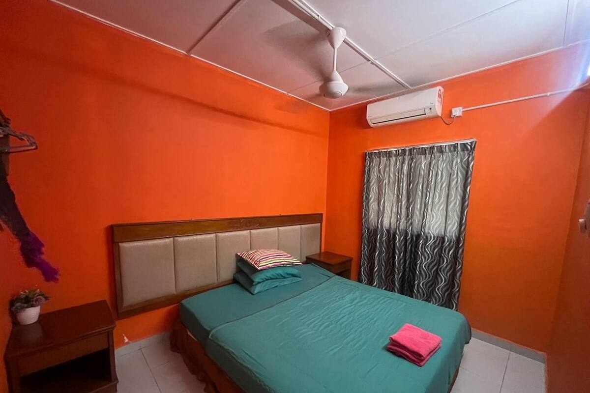 A bedroom with an orange wall, a wooden headboard, a teal bedspread, a pink towel, a ceiling fan, and an air conditioning unit.