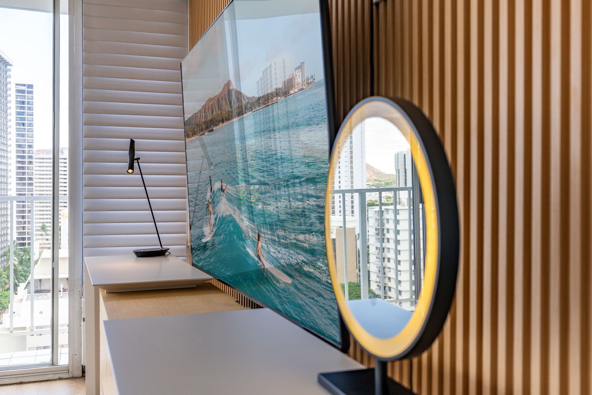 A room with a large flat-screen TV mounted on a wall, a round mirror on a stand, and a window with blinds.