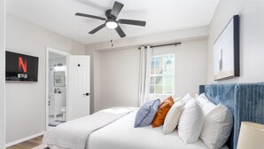 A bedroom with a bed, a ceiling fan, a TV mounted on the wall, a window with curtains, and a door leading to a bathroom.