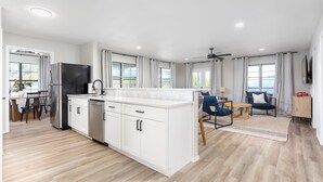A modern kitchen with white cabinetry, a stainless steel refrigerator, and a built-in oven. There is a dining area with a wooden table and chairs, and a living area with a sofa and a TV.