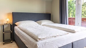 A double bed with a grey headboard, a bedside table with a lamp, and a view of greenery through a window. - Comfy Room in Shared Villa w/ Free Parking (Wien)