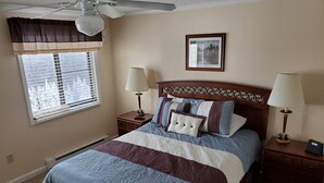 A bedroom with a bed, two bedside tables, a ceiling fan, a window with blinds, and a framed picture on the wall.
