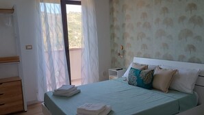 A bedroom with a bed, a wooden dresser, a nightstand, and a window with sheer curtains.