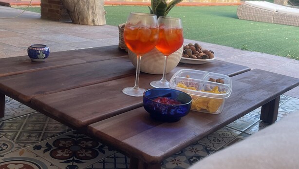 An outdoor dining area with a wooden table, a decorative plant, two glasses of red drink, and a bowl of snacks.