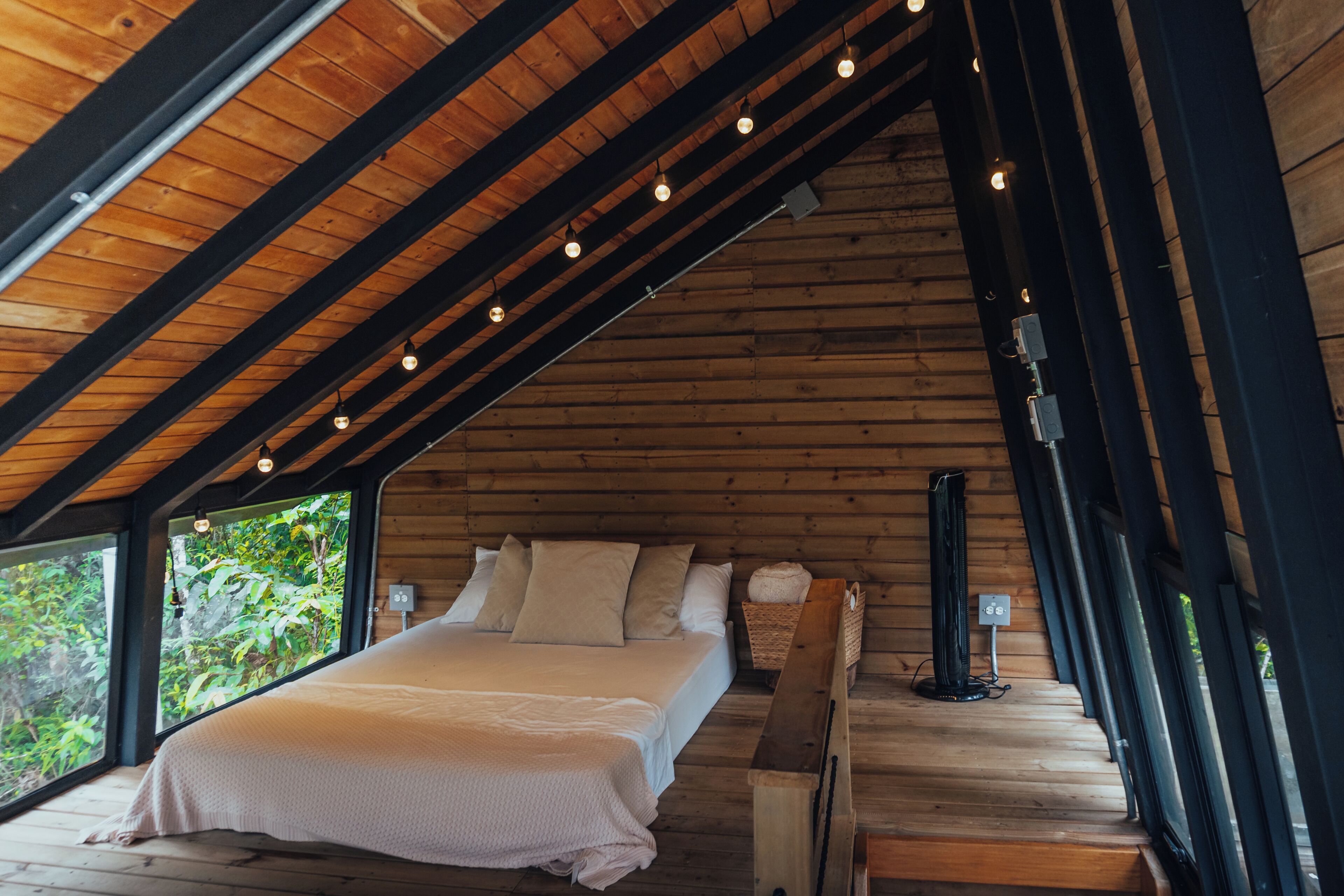 A wooden room with a bed, a bench, and a window overlooking greenery.
