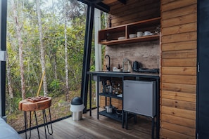 Eine kompakte KĂŒche mit einer HolzvertĂ€felung, einem kleinen KĂŒhlschrank und einem Fenster mit Blick auf einen Wald.