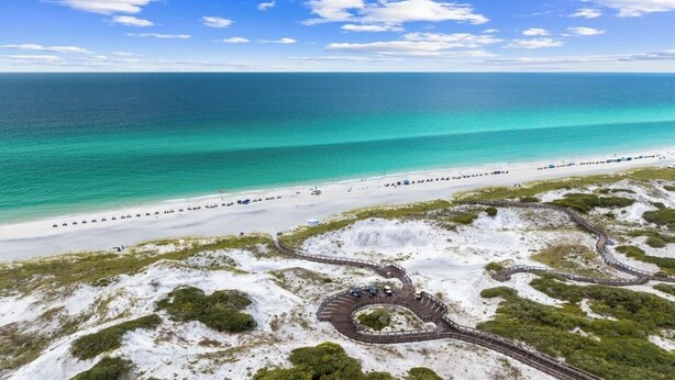 Vista aérea de una playa con un paseo marítimo serpenteante que conduce a un área circular de observación, rodeada de arena blanca y vegetación verde.