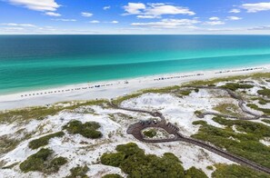 An aerial view of a beach with a winding boardwalk leading to a circular viewing area surrounded by white sand and green vegetation.