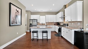 A modern kitchen with white cabinets, a large island with stools, stainless steel appliances, and a tiled backsplash. - Hill Country Retreat & Casita (Horseshoe Bay)