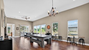 A spacious living area with a pool table, a ceiling fan, and large windows offering a view of the outdoors. - Hill Country Retreat & Casita (Horseshoe Bay)