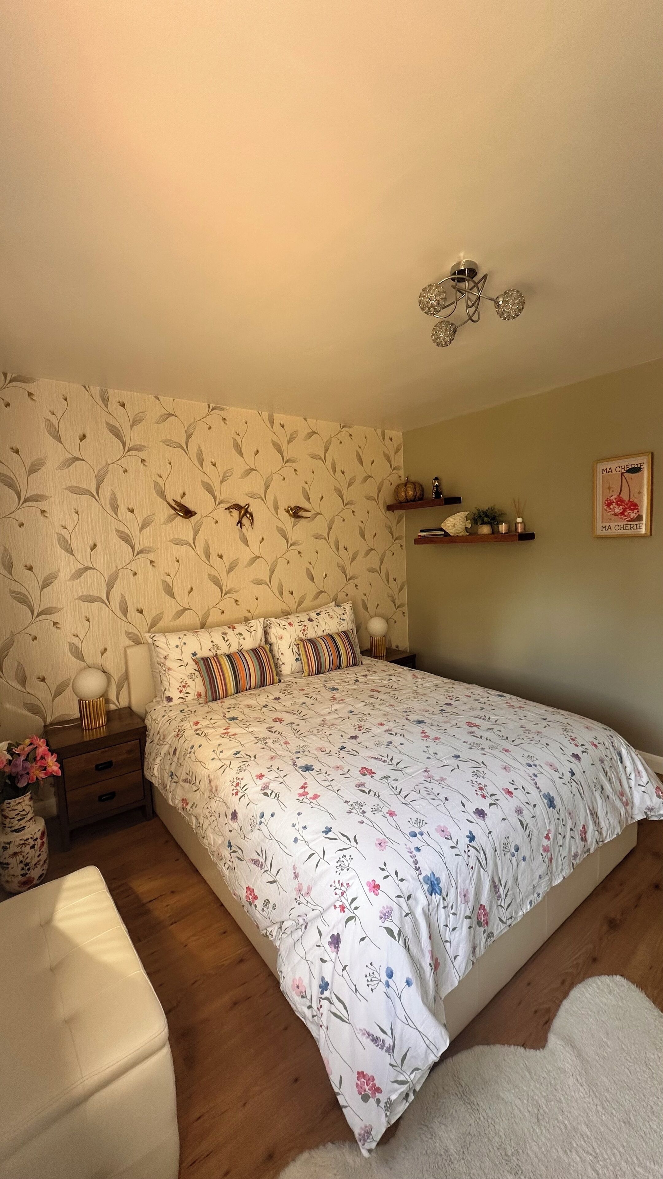 A bedroom with a floral-patterned bedspread, a nightstand with a lamp, a dresser, and a wall-mounted shelf with decorative items.