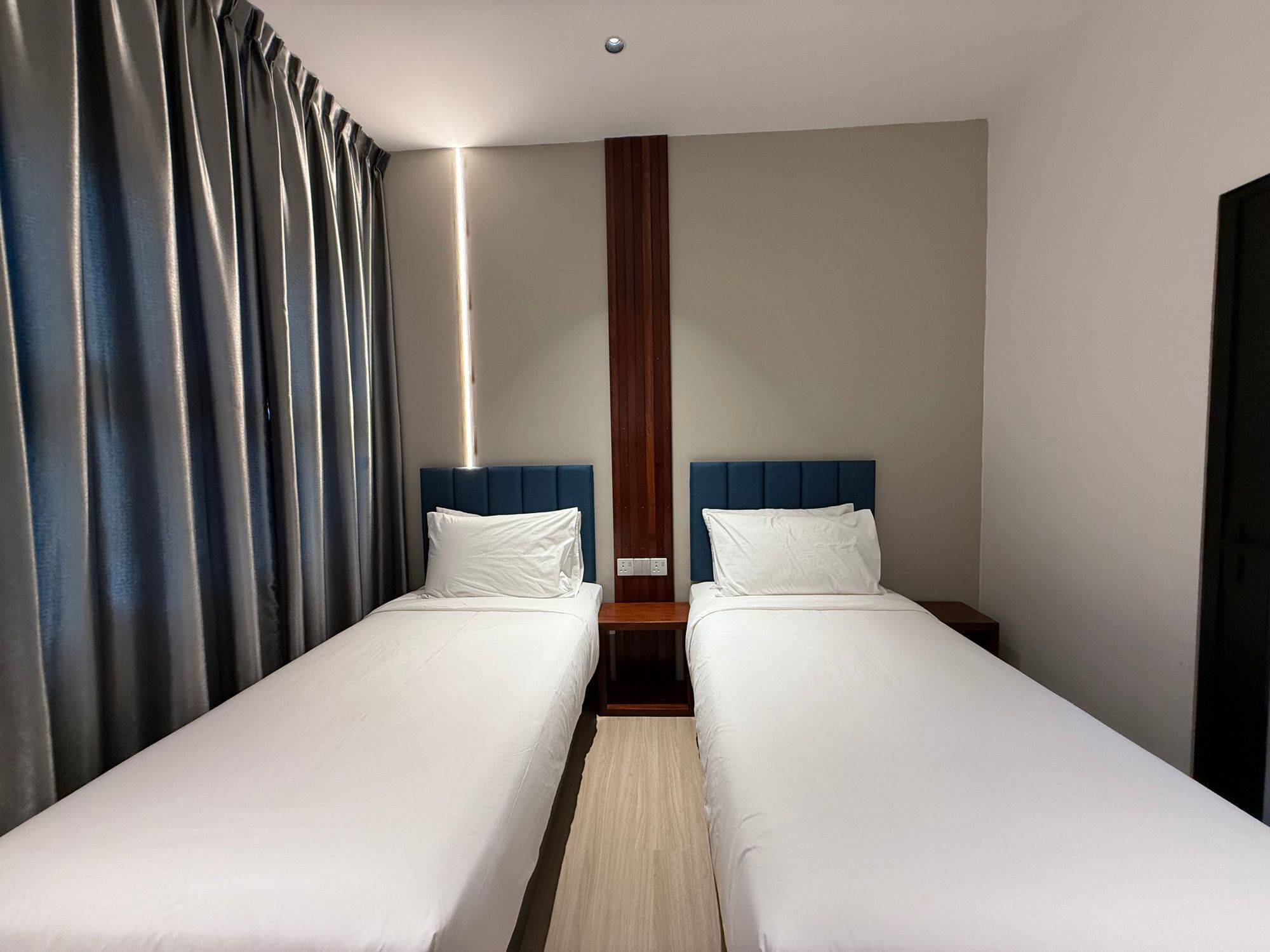 A hotel room with two single beds, a wooden nightstand, a modern light fixture, and a large window with curtains.