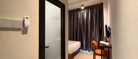 Habitación de hotel con una cama, un escritorio con una silla, una mesita con una botella y un cubo de basura.