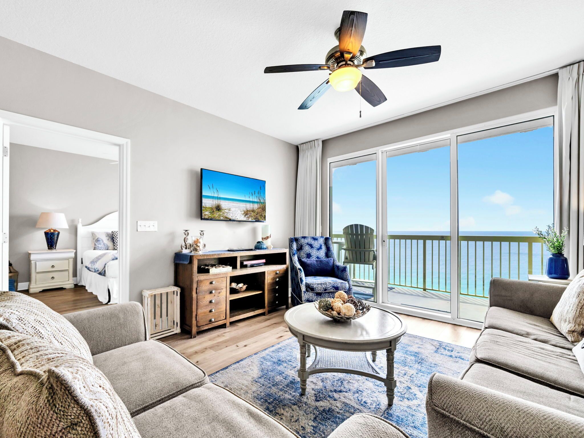 A spacious living room with a ceiling fan, a large window with a view of the ocean, a comfortable sofa set, and a small table with decorative items.