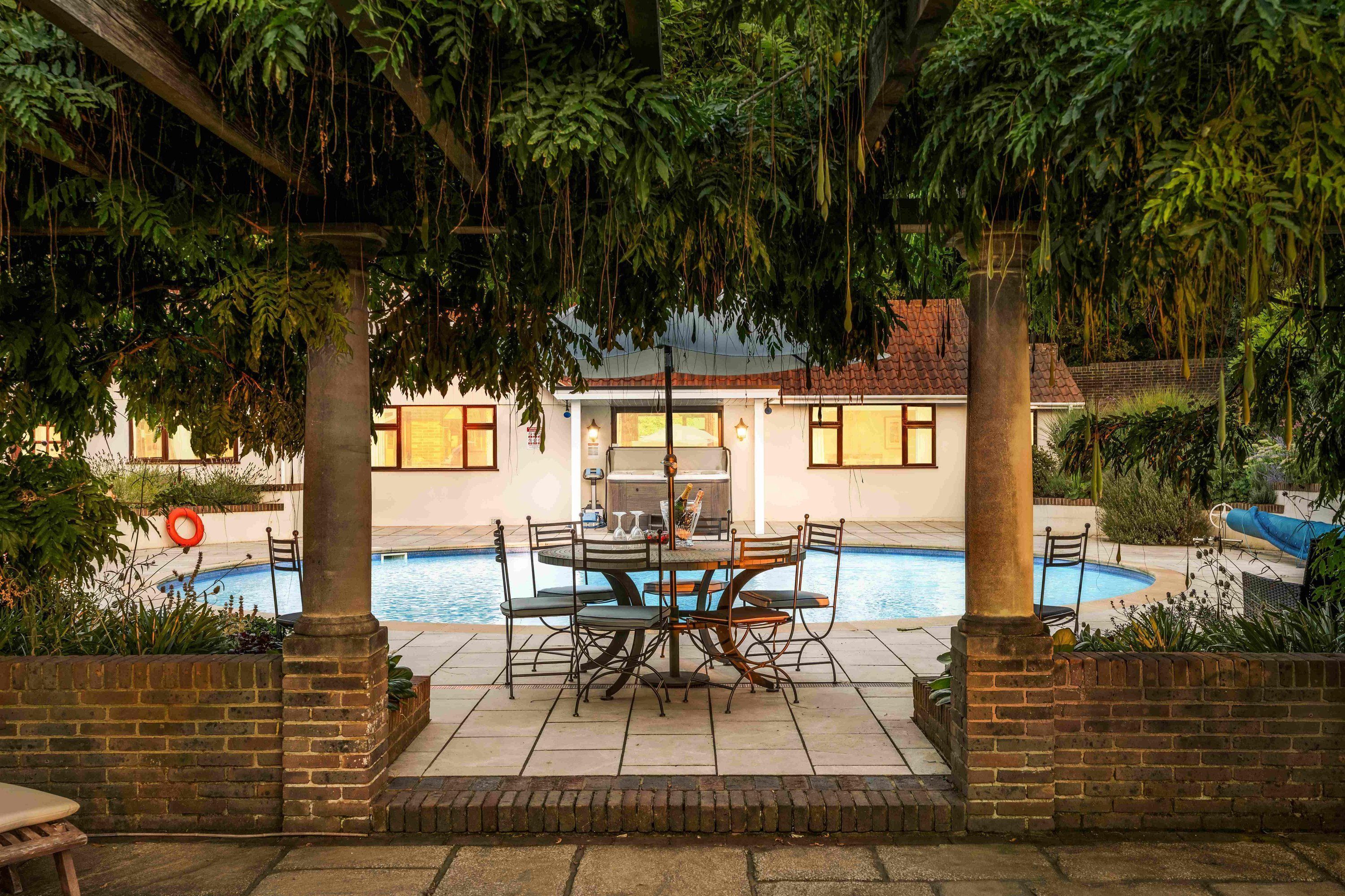A poolside area with a dining table and chairs, surrounded by lush greenery and a view of a building in the background.