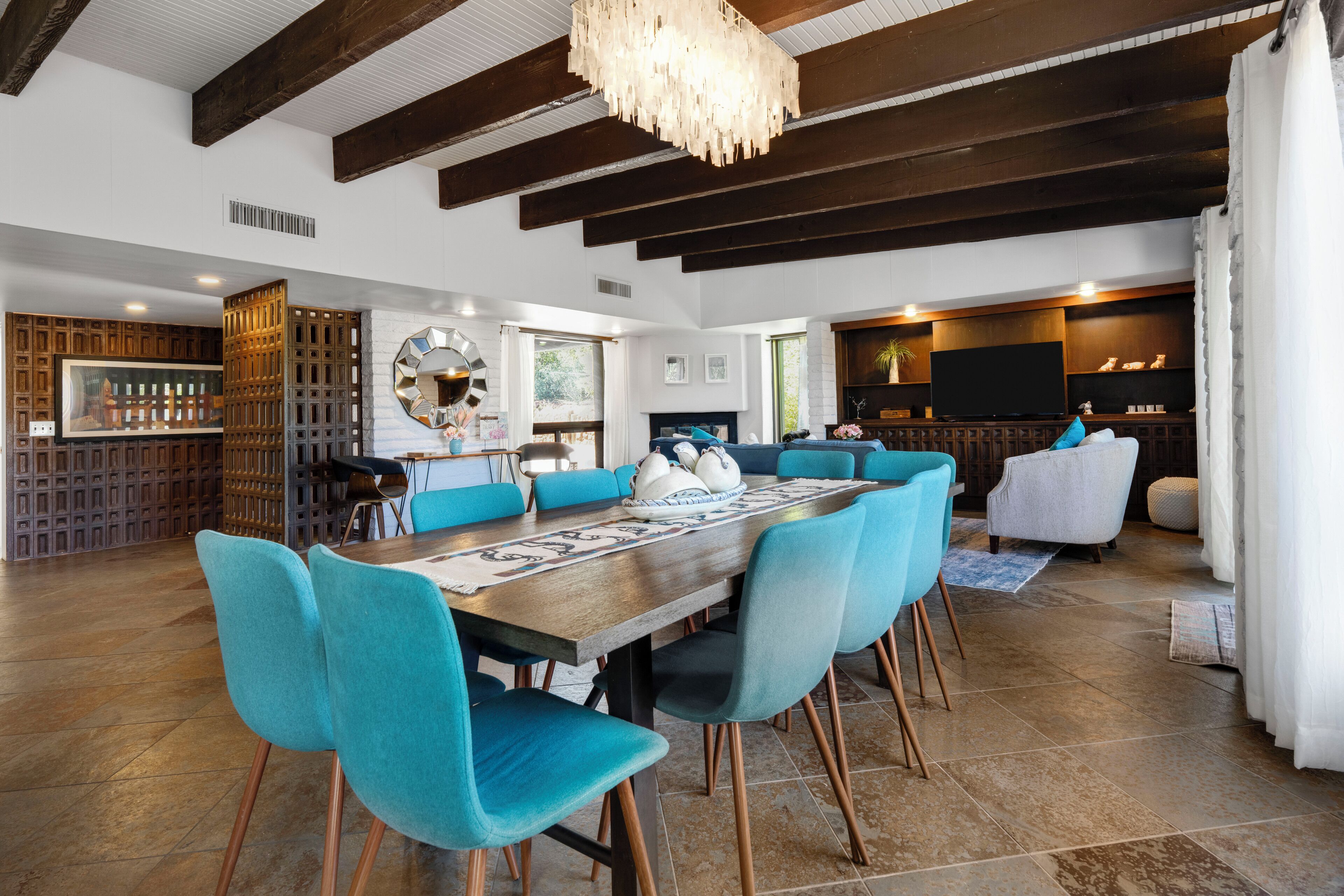 A spacious living area with a large wooden dining table surrounded by teal chairs, a fireplace, and a television in the background.