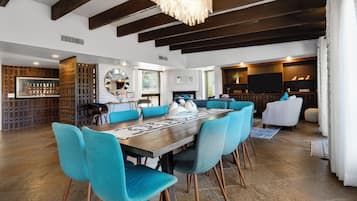 A spacious living area with a large wooden dining table surrounded by teal chairs, a fireplace, and a television in the background.