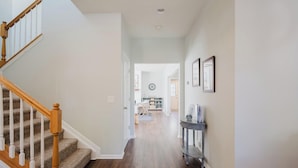 A hallway with a wooden staircase on the left, a console table with framed pictures on the right, and a view into a living area with a couch and a dining table. - Bluffton Magnolia House (Bluffton)