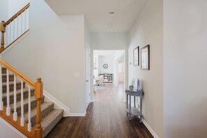 A hallway with a wooden staircase on the left, a console table with framed pictures on the right, and a view into a living area with a couch and a dining table. - Bluffton Magnolia House (Bluffton)