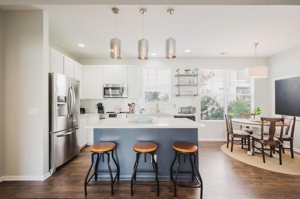 A modern kitchen with a central island, bar stools, and a dining area with a table and chairs. - Bluffton Magnolia House (Bluffton)
