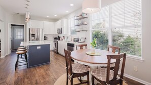 A modern kitchen with white cabinets, a central island, and a dining area with a round table and chairs. - Bluffton Magnolia House (Bluffton)