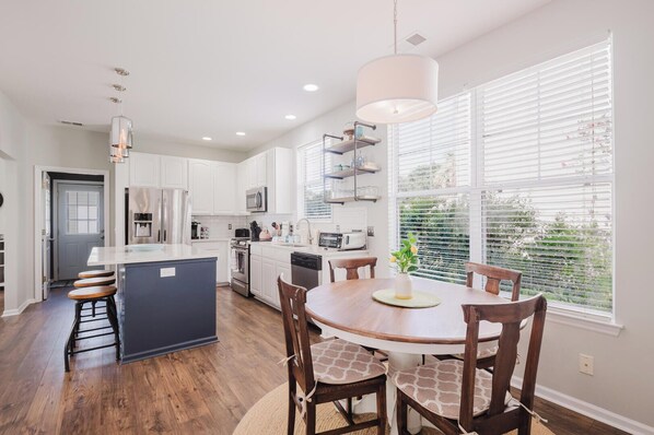 A modern kitchen with white cabinets, a central island, and a dining area with a round table and chairs. - Bluffton Magnolia House (Bluffton)