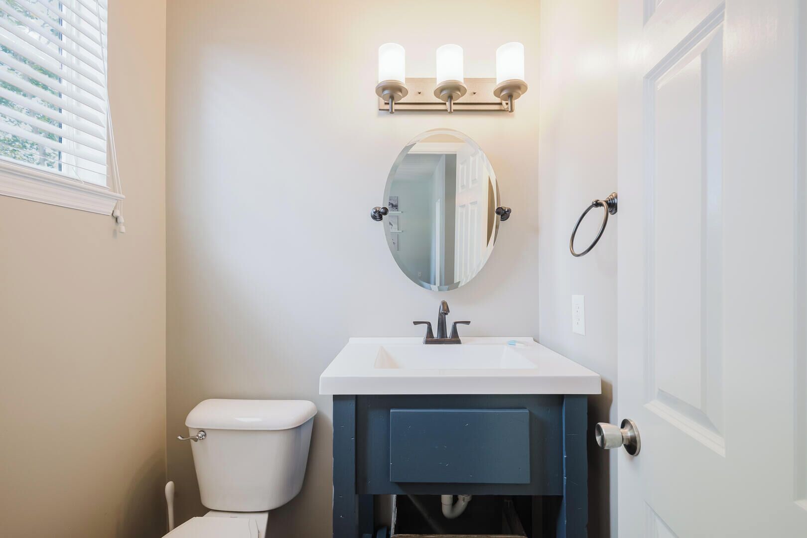 A bathroom with a white toilet, a white sink with a blue cabinet, a round mirror, and a wall-mounted light fixture.