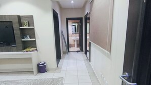 Un pasillo con piso de baldosas, un televisor sobre un pedestal, un cesto de basura y una puerta a la derecha.