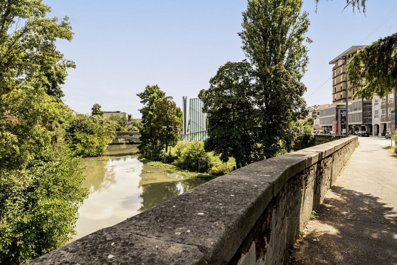 Un río con un puente de piedra, árboles y edificios al fondo.