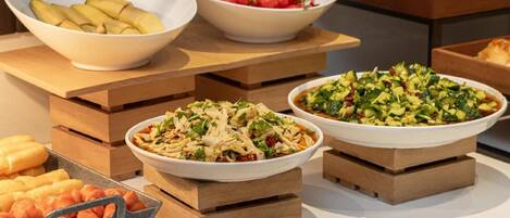 A buffet table with various dishes including sliced bananas, cherry tomatoes, and a salad.