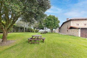 Un espacio al aire libre con césped, mesas de picnic, un edificio de piedra con tejado de tejas y árboles.