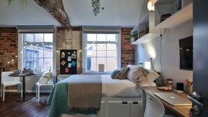 Une chambre avec un lit, un bureau, une chaise et un coin repas comprenant une table et des chaises. On y trouve des fenêtres, un mur en briques et une plante suspendue au plafond.