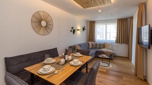 A modern living room with a dining area, a grey sofa, a wooden dining table set for four, and a television mounted on the wall.