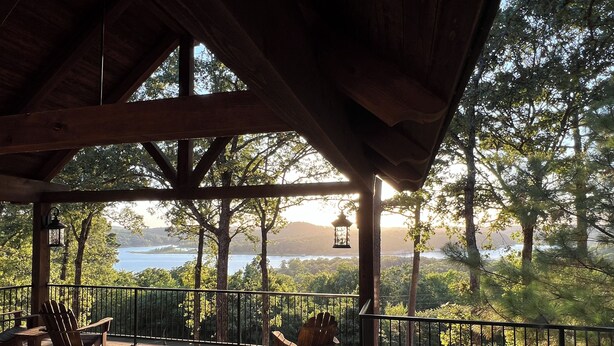 A covered deck with wooden furniture, a view of a lake, and trees in the background.