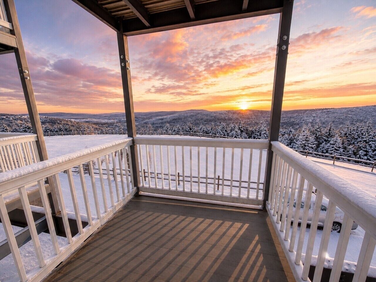 Un espace extérieur abrité, doté d’une terrasse en bois et de rampes blanches, qui offre une vue sur un paysage enneigé illuminé par le coucher du soleil.