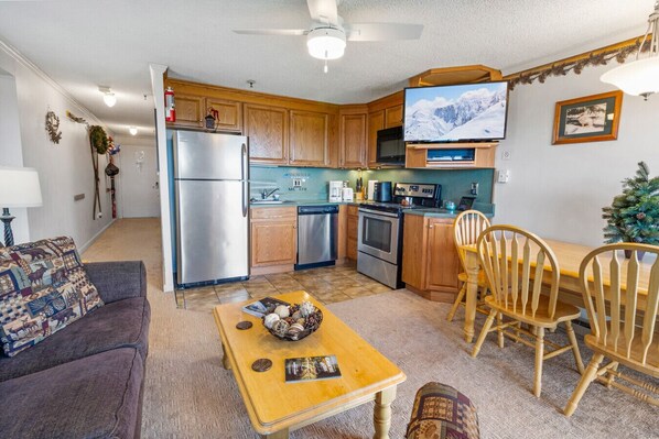 A compact kitchen with wooden cabinets, a refrigerator, microwave, stove, and a TV. There's a dining area with a table and chairs, and a small Christmas tree. - ML178 Great Views & Location EZ Parking  Next To Village (Snowshoe)