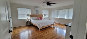 Quarto com piso de madeira, cama com roupa de cama branca, ventilador de teto e duas janelas com persianas.