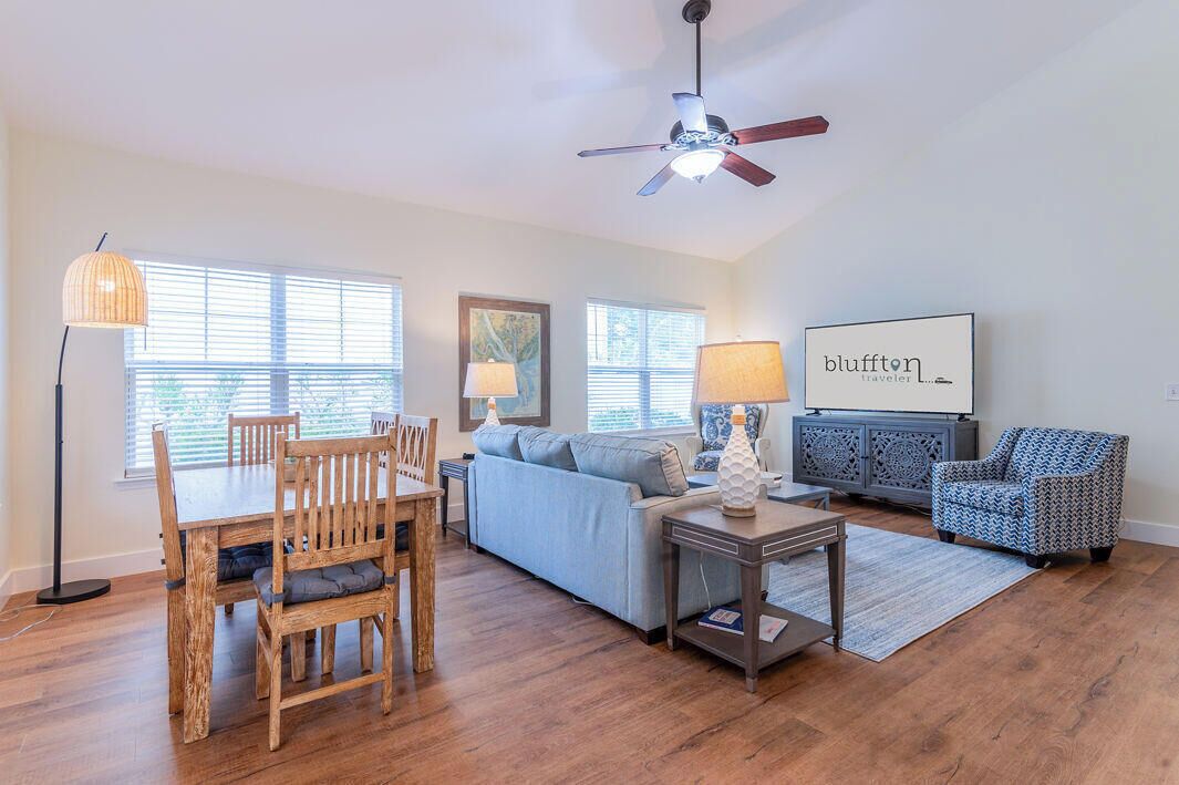 A living room with a dining table, chairs, a sofa, a patterned armchair, a ceiling fan, and a wall-mounted TV.