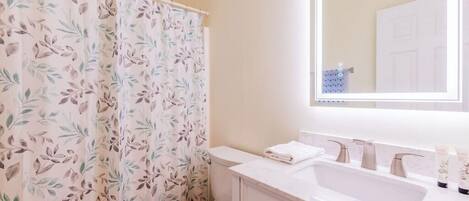 A bathroom with a floral shower curtain, a toilet, a sink with a mirror, and toiletries on the counter.