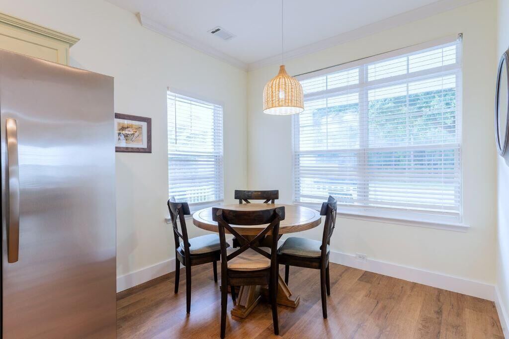 A dining area with a round table and four chairs, large windows with blinds, a hanging light fixture, and a refrigerator.