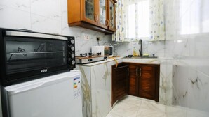 A compact kitchen with a built-in microwave, a refrigerator, wooden cabinets, and a window with floral curtains. - Entebbe Serenity breeze apartments - Single Room with Lake View (Entebbe)